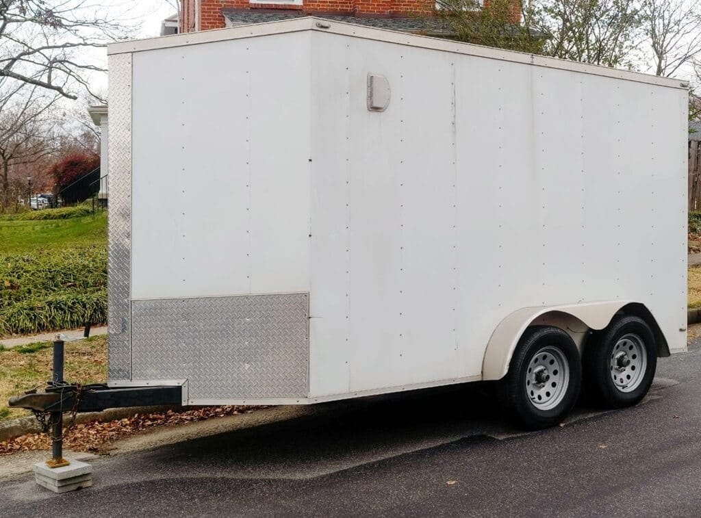 A white enclosed utility trailer with two axles and a silver diamond-plate panel on the front is parked on the side of a residential street